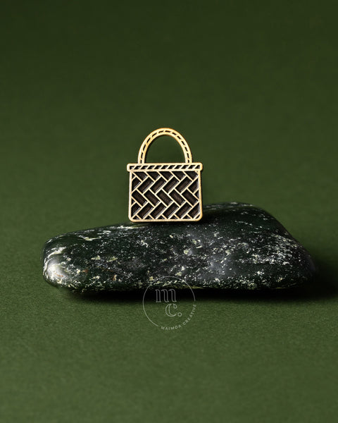 Gold woven kete basket pin on a dark greenstone pounamu surface with a green background