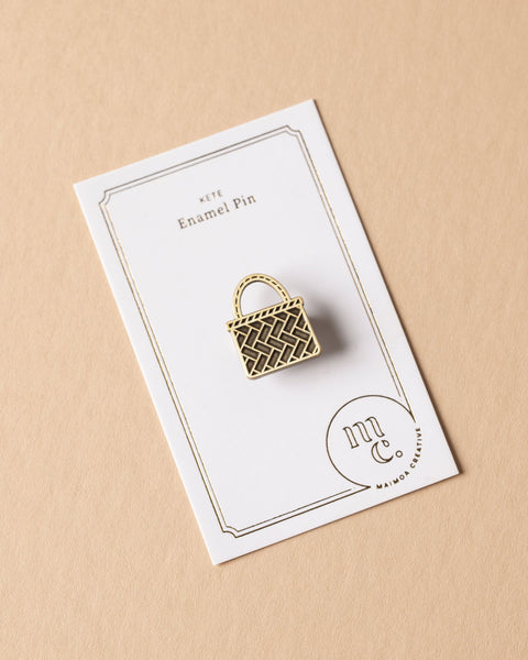Gold kete enamel pin on a white card with Maimoa Creative gold foil branding on a beige background