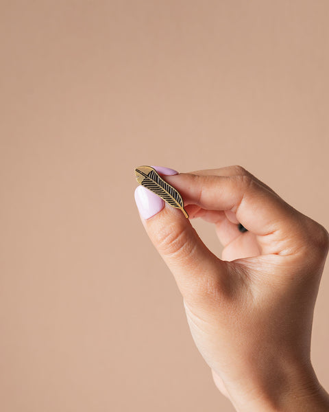 Hand holding a small gold huia feather pin against a beige background