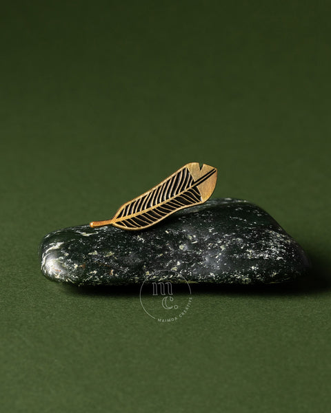 Gold huia feather brooch on a dark green stone pounamu against a green background