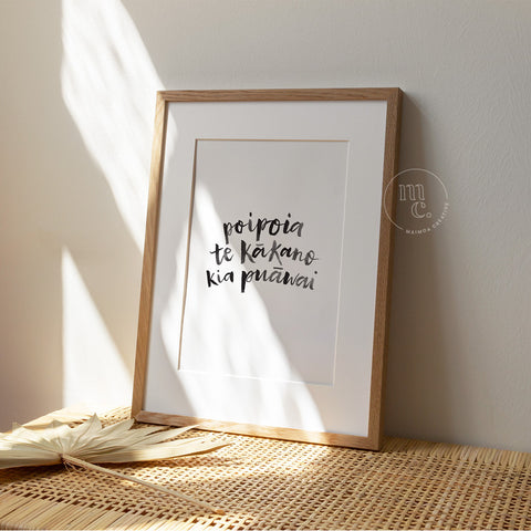 Framed A4 Māori art print with the quote “Poipoia te kākano kia puāwai” displayed in sunlight on a woven rattan surface.