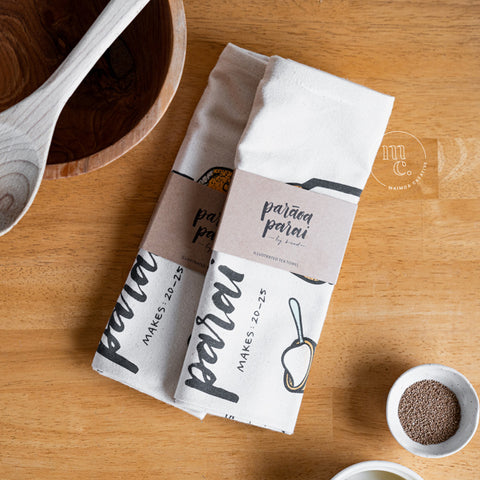 Two folded tea towels with an illustrated recipe for Māori fry bread, wrapped with a kraft paper band labeled 'parāoa parai', next to a wooden bowl and a small white bowl of yeast, on a wooden tabletop.