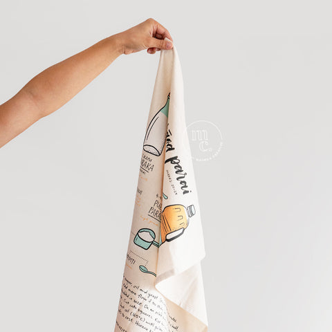 A hand holding up a corner of an illustrated tea towel featuring elements of a Māori fry bread recipe, with drawings of a glass bottle of milk, a cup of flour, oil, and other ingredients against a light background.
