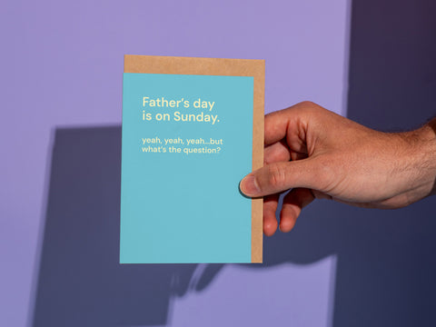 Hand holding a funny Father’s Day card on a purple background with the quote “Father’s day is on Sunday. yeah, yeah, yeah…but what’s the question?” by Maimoa Creative.