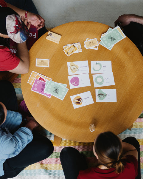 People sitting around a wooden table with reo māori cards from Tipu game spread out on a colourful rug.
