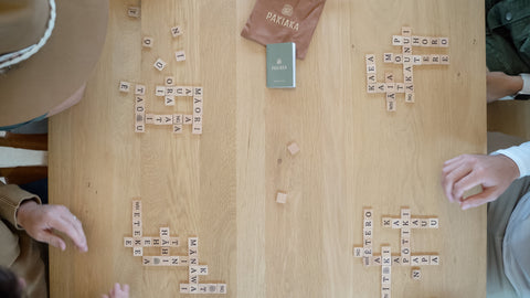 Overhead view of four people's hands reaching into a scattered pile of wooden letter tiles on a table as they engage in a word-forming Māori game.