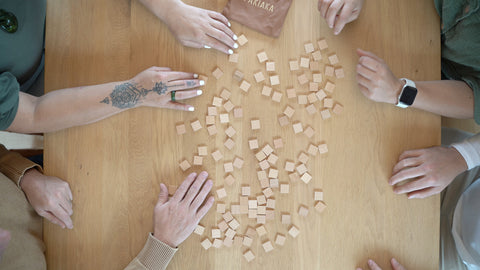 Overhead view of four people's hands reaching into a scattered pile of wooden letter tiles on a table as they engage in a word-forming Māori game.