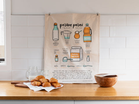 A kitchen setting with a tea towel hung on the wall featuring illustrations and recipe instructions for making frybread, accompanied by a plate of freshly made frybread on a wooden board, a mason jar, and a wooden bowl on a countertop.