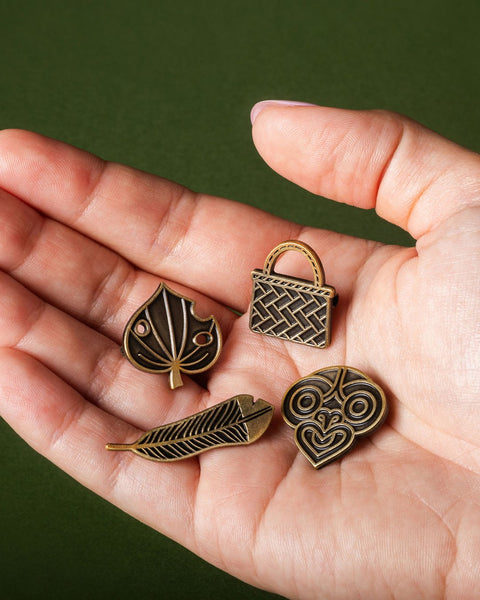 Hand holding four gold pins with different designs against a green background
