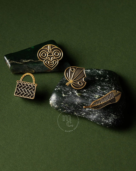 Gold pin collection with kete, tiki, kawakawa leaf and huia feather design on a dark green pounamu surface.