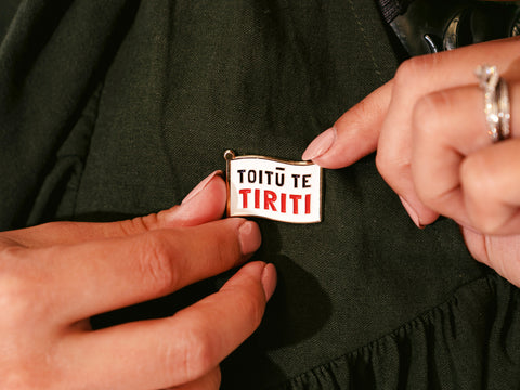 Two hands securing an enamel pin with the words "Guided By My Tūpuna" written in gold letters onto a dark forest green dress with another pin next to it saying "Toitū Te Tiriti"