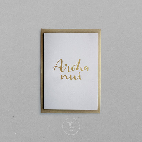 Flat lay of a premium reo Māori greeting card with 'Aroha nui' in gold foil lettering with gold envelope on plain background