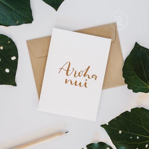 Flat lay of a premium reo Māori greeting card with 'Aroha nui' in gold foil lettering, gold envelope, surrounded by green kawakawa leaves and a pencil