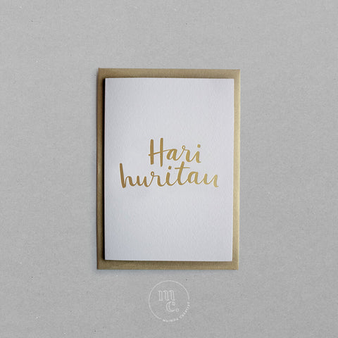 Flat lay of a premium reo Māori greeting card with 'Hari Huritau' in gold foil lettering with gold envelope on plain background