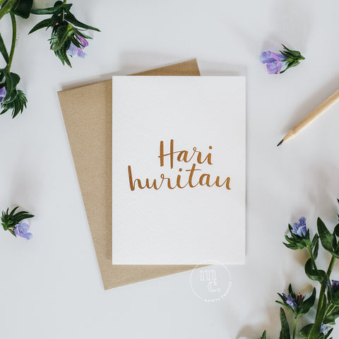 Flat lay of a premium reo Māori greeting card with 'Hari Huritau' in gold foil lettering, gold envelope, pencil, and scattered purple flowers.