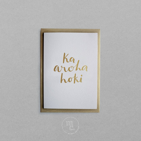 Flat lay of a premium reo Māori greeting card with 'Ka aroha hoki' in gold foil lettering and gold envelope on plain background