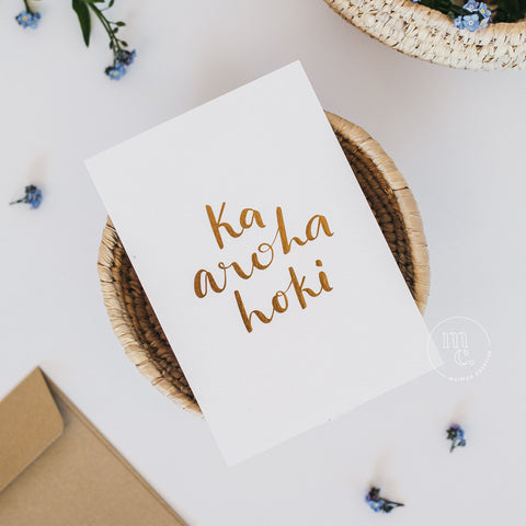 Flat lay of a premium reo Māori greeting card with 'Ka aroha hoki' in gold foil lettering and gold envelope with an earthy basket with purple scattered flowers surrounding it