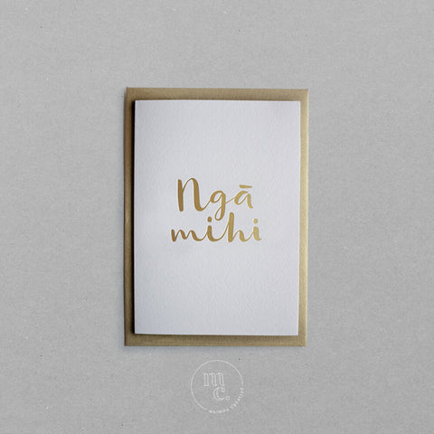 Flat lay of a premium reo Māori greeting card with 'Ngā Mihi' in gold foil lettering and gold envelope on plain background