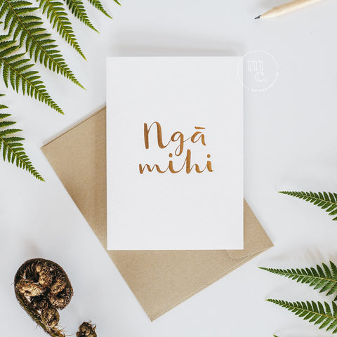 Flat lay of a premium reo Māori greeting card with 'Ngā Mihi' in gold foil lettering and gold envelope surrounded with green ferns, fern frond and pencil