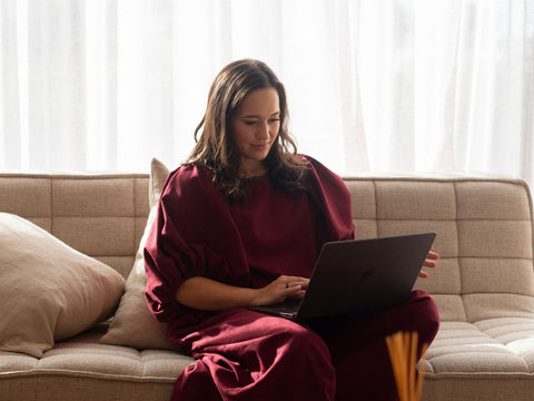 māori woman in a plum outfit using a laptop on a beige couch with white curtains in the background
