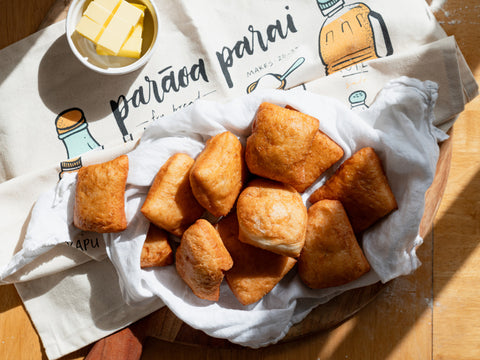 A pile of golden-brown Māori fry bread on a wooden table, wrapped in a white cloth, with a illustrated tea towel and ramekin of cubed butter and natural sunlight streaming in