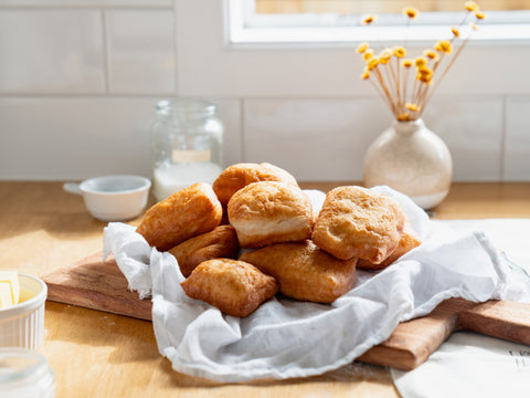 A pile of golden-brown Māori fry bread rests on a white cloth over a wooden cutting board, with kitchen items like a jar, a white ramekin, and a vase with dried yellow flowers in the background, all illuminated by natural light on a wooden countertop.