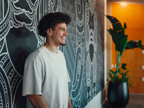 Māori man with afro smiling wearing white t-shirt in front of large Māori mural sharing his pepeha