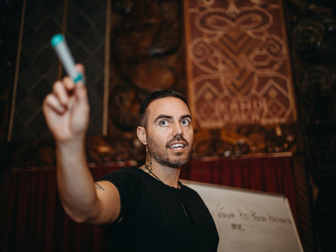 A Māori educator in a traditional wharenui holds up a marker while teaching, with intricate Māori carvings in the background and a whiteboard with te reo Māori words written on it