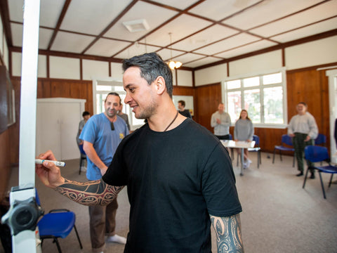 A Māori man with traditional tattoos writing on a whiteboard in a class with adult learners smiling in the background.