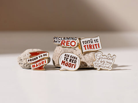 A collection of five enamel badge pins promoting te reo Māori, including 'Reclaiming My Reo', 'Toitū te Tiriti', 'Proud to be Māori', and 'Guided by My Tūpuna', displayed on natural pumice stone with clear details and brass accents.