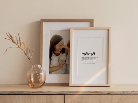 Two framed prints on a wooden shelf; on the left, a mother-daughter photo, and on the right, a 'māmā' definition print, with a vase and dried flowers beside them.