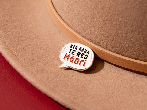 Close-up of a 'Kia Kaha Te Reo Māori' enamel pin attached to the band of a tan felt hat, symbolizing support for the strength of the Māori language.