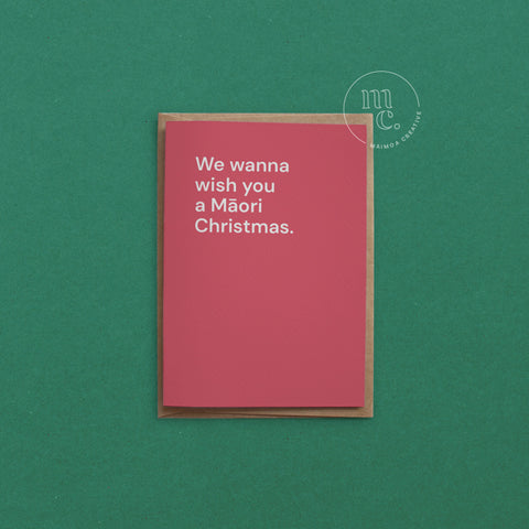 Greeting card with text 'We wanna wish you a Māori Christmas' on a bright red background, placed on a green surface