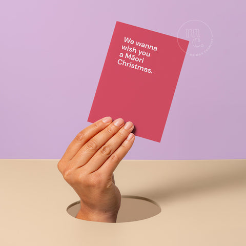 Hand peeking out of hole holding a Christmas greeting card with the text 'We wanna wish you a Māori Christmas' against a pastel purple and beige background.
