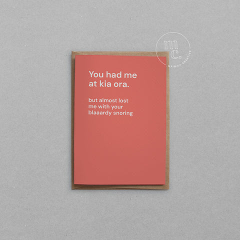 Greeting card with text 'You had me at kia ora but almost lost me with your blaaardy snoring' on a bright peach background, placed on a light surface