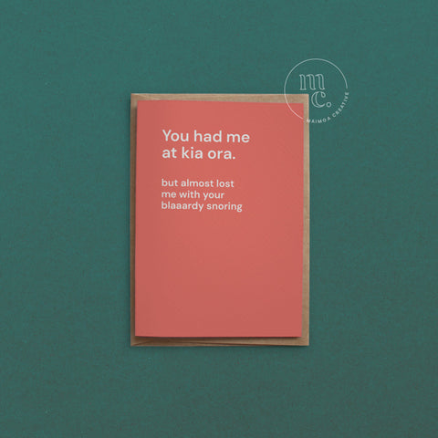 Greeting card with text 'You had me at kia ora but almost lost me with your blaaardy snoring' on a bright peach background, placed on a contrasting surface