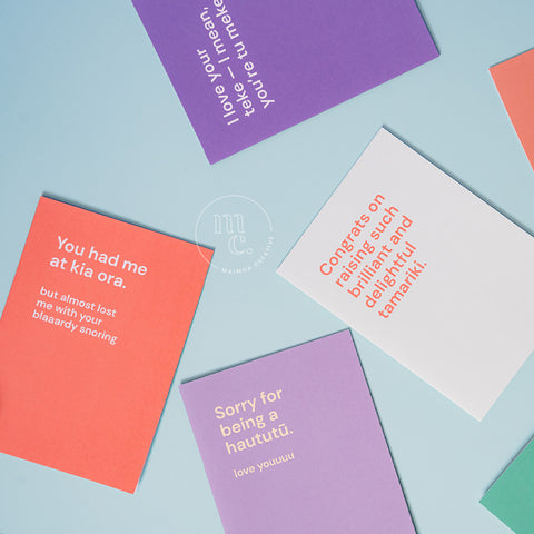 Assorted colourful cheeky greeting cards with playful Māori-themed messages spread on a pastel blue background.