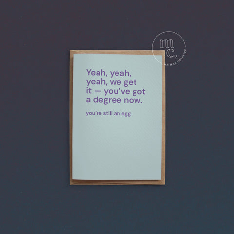 Greeting card with text 'Yeah, yeah, yeah, we get it — you’ve got a degree now. You’re still an egg' on a bright pastel blue background, placed on a contrasting surface