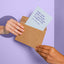 Two hands passing and receiving a humorous graduation greeting card poking out of a kraft envelope, with the message 'Yeah yeah yeah we get it—you’ve got a degree now,' on a pastel purple background
