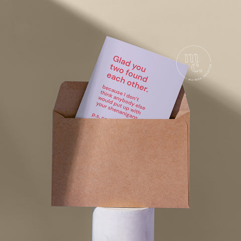 Cheeky card peeping from a kraft envelope on a plinth with the text 'Glad you two found each other' against a beige background.