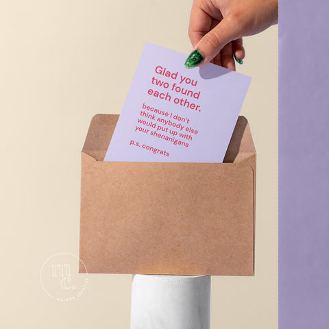 Cheeky card being pulled by a hand from a kraft envelope on a plinth with the text 'Glad you two found each other. Because I don't think anybody else would put up with your shenanigans,' against a pastel cream background.