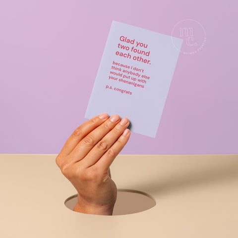 Hand peeking out of hole holding a wedding greeting card with the text 'Glad you two found each other. Because I don't think anybody else would put up with your shenanigans,'against a pastel purple and beige background.