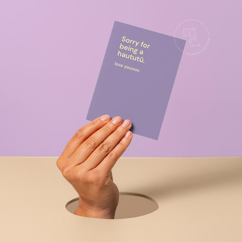 Hand peeking out of hole holding a greeting card with the text 'Sorry for being a haututū. love youuuu,' against a pastel purple and beige background.