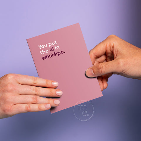 Hand passing an R18 greeting card with the text 'You put the ai in whaiāipo' on a pastel purple background