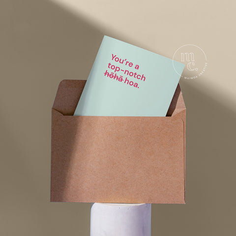 Cheeky card peeping from a kraft envelope on a plinth with the text 'You’re a top-notch hōhā / hoa.' against a beige background.