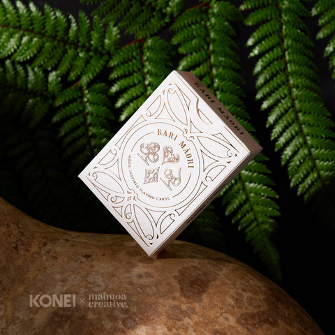 Kāri Māori playing cards balancing on a natural hue māori gourd, set against lush ferns in a moody, dark setting.