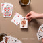 Hand revealing a Jack of diamonds playing card with a unique Māori-inspired design from a deck featuring Māori illustrations, with more cards scattered in the background