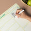 Hand writing a grocery list on a green magnetic weekly planner with a marker, next to a bowl of avocados and green apples