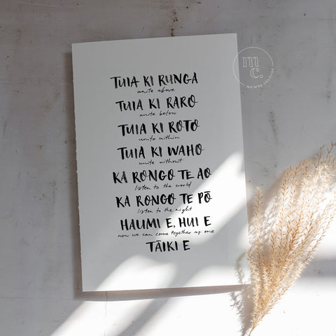 Print of an opening karakia print 'Tuia ki runga' with translation by Maimoa Creative on plain background with sunlight and plant.