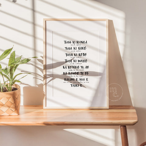 Framed opening karakia print 'Tuia ki runga' by Maimoa Creative on wooden shelf with sunlight and plant.